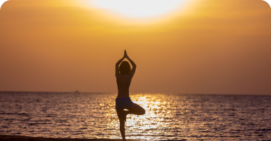 imagem de um por do sol na praia com uma mulher em pé realizando uma posição de yoga.