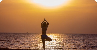 imagem de um por do sol na praia com uma mulher em pé realizando uma posição de yoga.